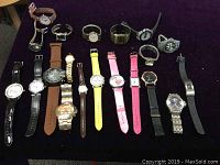 Wide overview of all 20 assorted quartz watches laid out on purple fabric surface, featuring a variety of strap styles and colors.