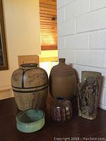 Five pottery items displayed on a wooden surface against a white brick wall, consisting of varied vessels and one sculpture.