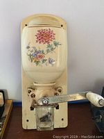 Front view of vintage cream ceramic coffee grinder with floral decoration and white handle mounted on cream backplate.