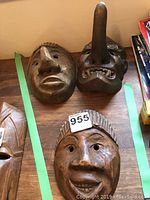 Photo showing three dark brown carved wood African masks on a table, two with neutral faces and one with a long nose and fanged mouth, tagged with lot number.