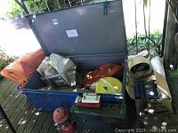 Blue steel lid-open trunk filled with tarps, outdoor gear, and a green Coleman stove base, with a red lantern and a red white Hunter flashlight outside in front