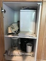 Photo showing inside kitchen cabinet with Black & Decker bread maker on upper shelf and Braun hand mixer with blender jug and espresso maker below.