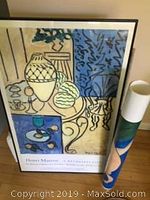 Framed Henri Matisse 'Still Life' poster featuring abstract shapes and vases with a mix of blue, cream, and black colors. Framed in black, shown standing on floor beside a rolled mat.