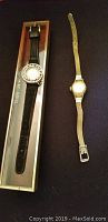 Two ladies wrist watches placed side by side. One has a leather strap in a box with 14K marking. The other has a gold-colored mesh strap.