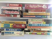 Photo showing an assortment of boxed board games stacked, including Password, Quilting Bee, Jeopardy, Superfection, Stratego, Parchessi, Yahtzee, Game of the States, Clue, Match Game, Rack-O, Numbers Up, Payday, and craft kits.