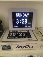Front view of DayClox large-font electric calendar clock displaying Sunday 3:29 PM, June 23, 2019, resting on original box