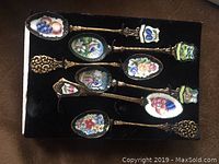 Top view of six cloisonné spoons showing detailed enamel floral and fruit patterns with ornate gold-tone handles, arranged on black fabric.