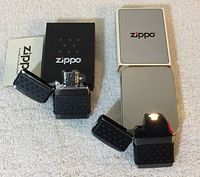 Photo showing both Zippo items open, displaying the lighter's chrome flame area and Ziplight with illuminated lamp, both on white fabric background alongside their boxes and manuals.