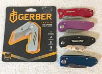 Five utility knives including a new Gerber E.A.B Lite in packaging and four others including Snap On and Trades Pro on a white towel.