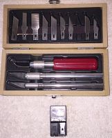 Open wooden box displaying three knives with red handles and multiple X-Acto blades arranged above in foam slots along with the sharpener/refill box below