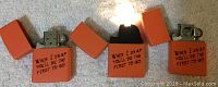 Photo showing three orange Zippo lighters with flip-top lids open exposing the metallic lighter insert. Engraved text on front: 'When I snap you'll be the first to go.'
