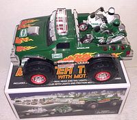 Green Hess monster truck toy in original box, showing flame decals, red rims, and motorcycles in truck bed