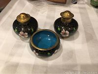 Three cloisonne items on white cloth: two salt and pepper shakers with brass tops and one small open container. Floral decorations with pink flowers, green leaves on black background visible. The inside of the open container shows turquoise blue.