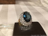Cocktail ring with large blue oval stone and clear rhinestones on a goldtone band, shown from an angled side view.