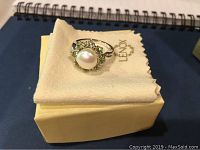 View of ring from top showing large pearl surrounded by light green stones, on Lenox branded cloth and box