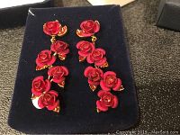 Pair of earrings shown from the front, each composed of five red roses vertically connected with gold-tone edges and leaves on a black display pouch.