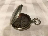 Silver tone metal pocket compass with hinged lid slightly open, showing white dial and compass needle.