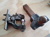 Photo showing two assorted trailer hitch couplers on a wooden surface, one with a square shank and hitch ball, the other with a mounting plate and hitch ball, both used and rusty.