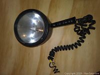 Photo of black MaxMillion 12V spot light with handle and coiled power cord on wooden surface.