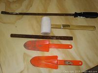 Photo showing assorted tools including two pry bars, a rubber mallet, a rusty metal rod, and two orange plastic garden trowels.