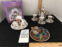 Three sets of miniature tea sets displayed against a black background with rulers for scale: a white porcelain set with red pomegranate and floral patterns, a purple floral tea set with gold accents, and a richly decorated brown teapot with small blue cups on a tray.