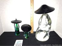 Three glass statues with black hats on a black surface against white background with measuring ruler and maxsold card
