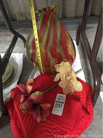 Image showing the red, green, and yellow glass vase with a measuring tape indicating height and two artificial flowers placed adjacent to it, along with red cloth textiles beneath.