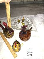 Photo showing two large swirl glass rooster figurines and three small multicolored glass roosters on a clear glass egg tray with a ruler for scale.
