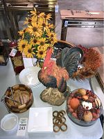 Rooster figurine, yellow artificial flowers, wooden bowl with salad tongs, white bowls and plates, glass bowl with artificial fruit, wooden napkin rings, marshmallow container, bottle of red liquid