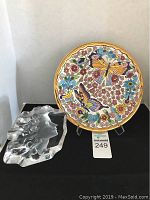 Photo showing crystalline sculpture of a lady's head next to a colorful butterfly and floral plate on stand, with MaxSold lot tag 249 visible.
