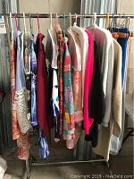 Photo of ladies tops and blouses hanging on a rack, showing variety of patterns and colors including pink, blue, cream, floral, and lace detail.