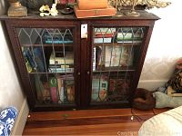 Front view showing dark wooden bookshelf with leaded glass doors filled with books. Glass on one door is broken.