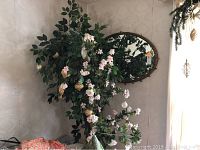 Wall mirror mounted on wall reflecting large leafy faux plant with pink flowers and ornaments hanging from it.