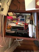 Top view of open desk drawer showing black Belkin desktop hub, scissors, stapler, tape, pens, adhesives, paper clips, envelopes, and note pads organized in compartments.