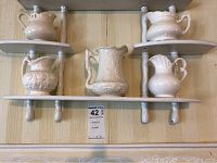 Close-up of one tier of the white wooden shelf with six cream-colored creamer pitchers showing details of design and paint chipping on shelf.