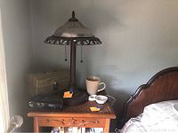 Metal table lamp with glass shade on wooden side table, cloth-covered box and black clock radio beside lamp