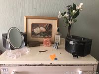 Overview of the items on a white dresser including jewelry box, vase with flowers, framed magnolia picture, animated salt candle holder, mirrors, and small digital device