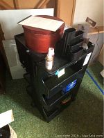 Black plastic art cart with multiple shelves, top shelf holding a red round container with paint tubes and various painting supplies including sprays and small bottles.