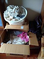 Laundry bin filled with white and neutral fabrics plus a cardboard box opened with mixed fabrics inside.