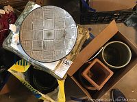 Photo of various ceramic pots and plates arranged on and near a wood side table, showing ceramic pots in different shapes and colors (beige, orange-brown, green) and a beige ceramic plate with a square pattern.