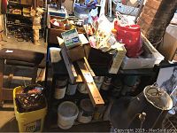 Photo of the basement table filled with assorted tools and items such as glaze compound containers, paint cans, a wooden miter box, spirit level, and assorted boxes with hardware and craft supplies.