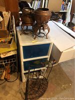Copper pots with spigots on stands placed on top of a white plastic organizer with blue drawers, set in a basement area