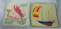 Two 13x13 inch needlepoint pillows together on white surface; one features a sailboat, the other a seashell design.