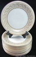 Photo showing entire stack of 10 salad plates with one plate on stand displaying ornate gold floral and scroll border on white porcelain base.