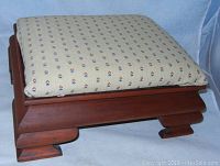 Front diagonal view of a vintage wooden footstool with a beige fabric cushion having a small multicolor pattern.