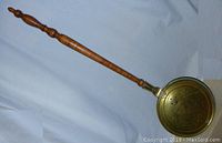 Full length view of the brass bed warmer showing the turned wood handle and attached brass bowl end resting on a white surface