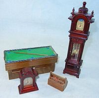 Four miniature dollhouse items: a tall grandfather clock, a mantel clock, a pool table with one cue stick, and a small wooden toolbox.