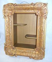 Front view of the ornate framed shadow box wall shelf showing detailed gold-colored frame with intricate patterns and two interior shelves inside the rectangular opening.