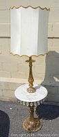 Full view of the lamp showing the sculpted gold-toned metal base, round white marble table surface with hanging crystal accents, and white scalloped lampshade with gold trim.