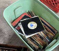 Top view of green plastic container filled with numerous 45 RPM vinyl records in sleeves showing random labels and covers.